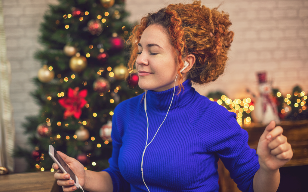 Kvinna i blå tröja har hörlurar och lyssnar på musik framför julgran.