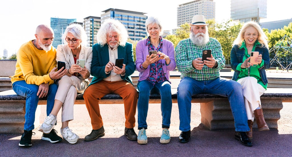 Äldre vänner sitter på bänk och använder mobiltelefoner.