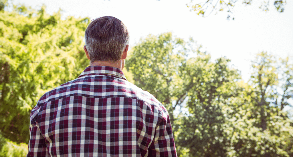 Man i rutig skjorta står med ryggen mot och tittar ut över gröna träd.